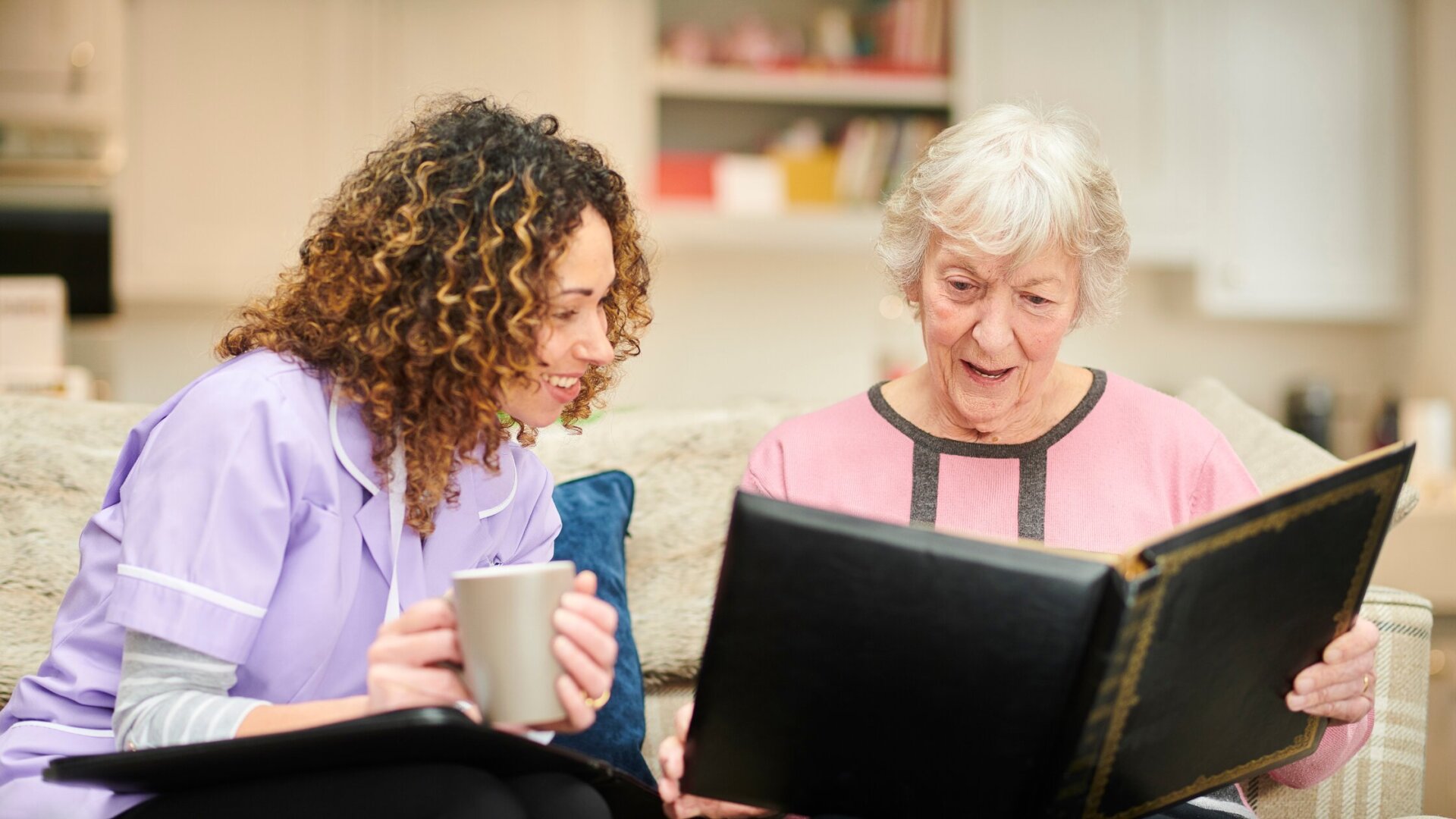 Senior woman sitting on the couch with her caregiver while looking through old photos at Traditions of Aboite